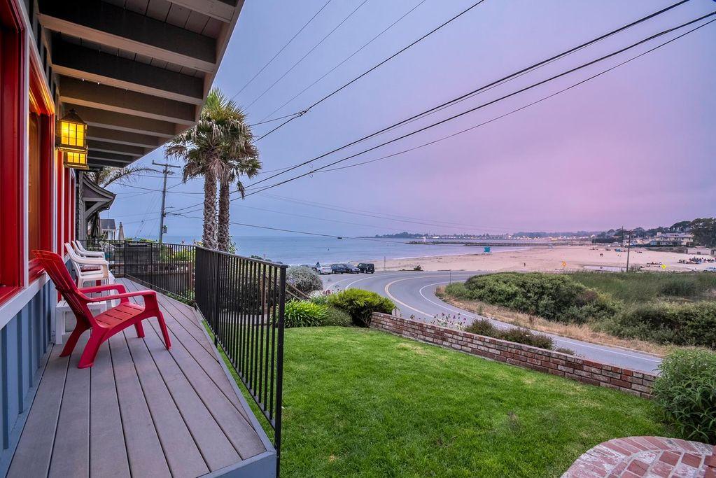 Charming beachfront bungalow in Santa Cruz with a surfboard on the porch, coastal landscaping, and a wooden walkway leading to the ocean, overlooking waves and nearby shoreline homes, capturing the laid-back surf lifestyle and prime oceanfront real estate in Santa Cruz California.