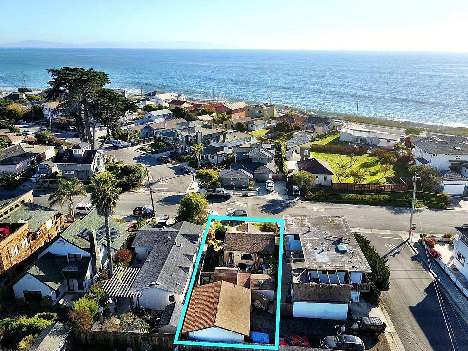 Ocean-view coastal home in Santa Cruz County featuring a large wooden deck overlooking Monterey Bay, surrounded by redwood trees and landscaped greenery, with a “For Sale” sign visible, showcasing desirable Santa Cruz real estate with outdoor living space and scenic ocean views.