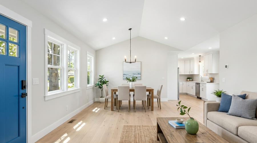 A bright open concept interior featuring fresh light neutral paint, clean wide plank wood flooring, a staged dining set with an olive branch centerpiece, and a vibrant blue front door letting in natural sunlight with zero clutter.