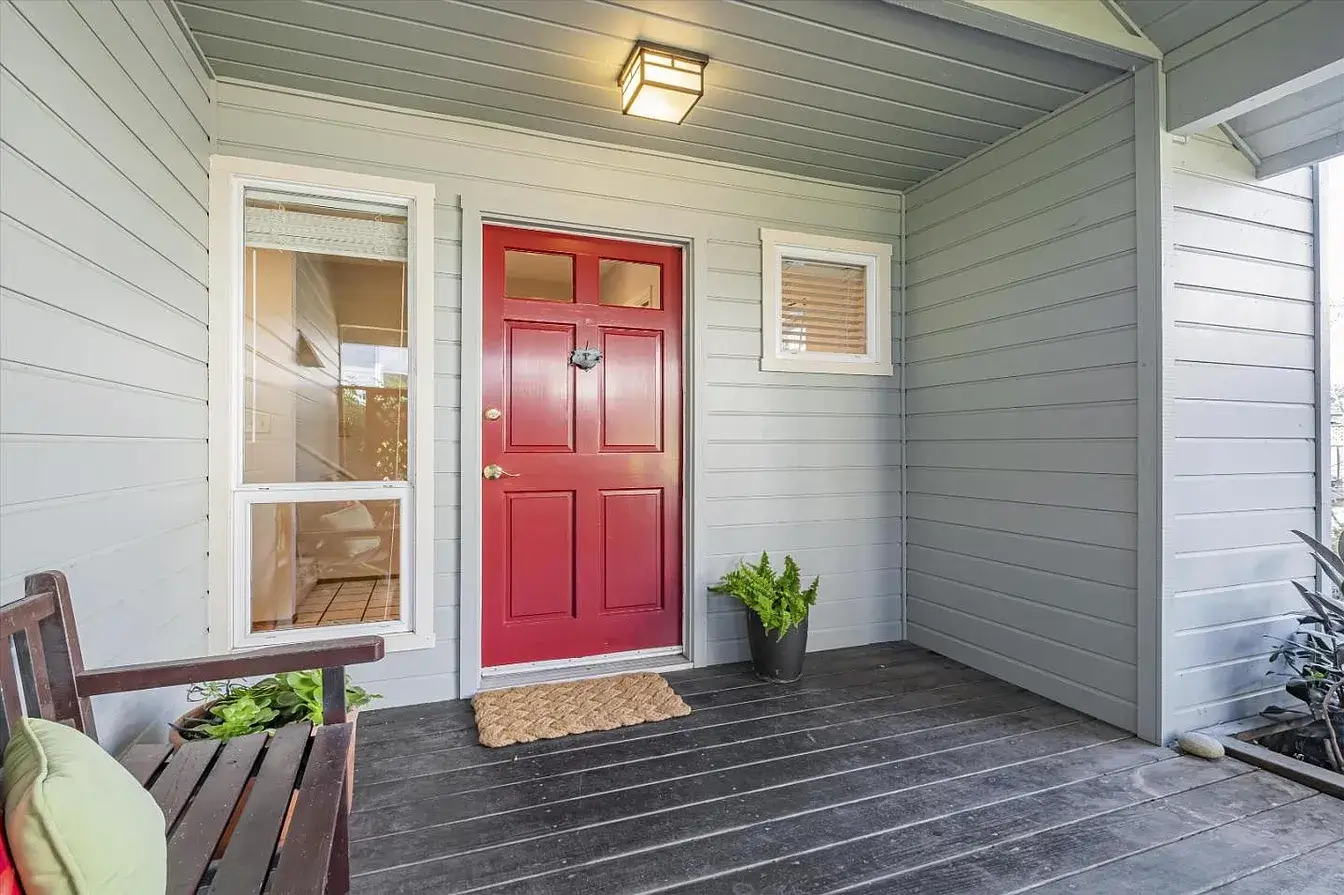 Covered front porch of a Santa Cruz home with gray exterior siding, a red front door, black-painted wood deck flooring, white trim, a porch bench, potted plants, and a simple entry light. This Santa Cruz property entry highlights clean curb appeal, inviting front porch design, and well-kept exterior details that support strong presentation in the Santa Cruz real estate market.
