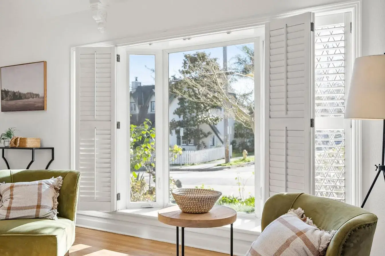 Bright and airy living room in a Santa Cruz home featuring a large bay window with white shutters, natural sunlight, two green accent chairs with patterned throw pillows, a small round wooden table with a wicker basket, light hardwood floors, and minimal decorative touches. This staged Santa Cruz property interior highlights comfort, curb appeal from inside, and a move-in ready presentation for homes for sale in Santa Cruz County.