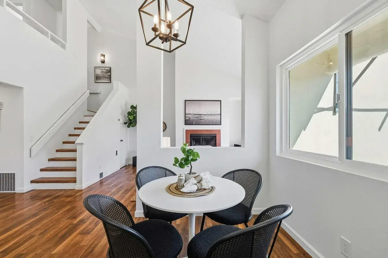 Staged Santa Cruz Dining Area With Staircase and High Ceilings Bright staged dining area inside a Santa Cruz home with a round white table, four black woven chairs, warm wood floors, tall ceilings, a modern pendant light, and an open staircase leading to the upper level. This Santa Cruz property interior highlights clean staging, natural light, and an open layout that can appeal to buyers comparing homes for sale in Santa Cruz County.