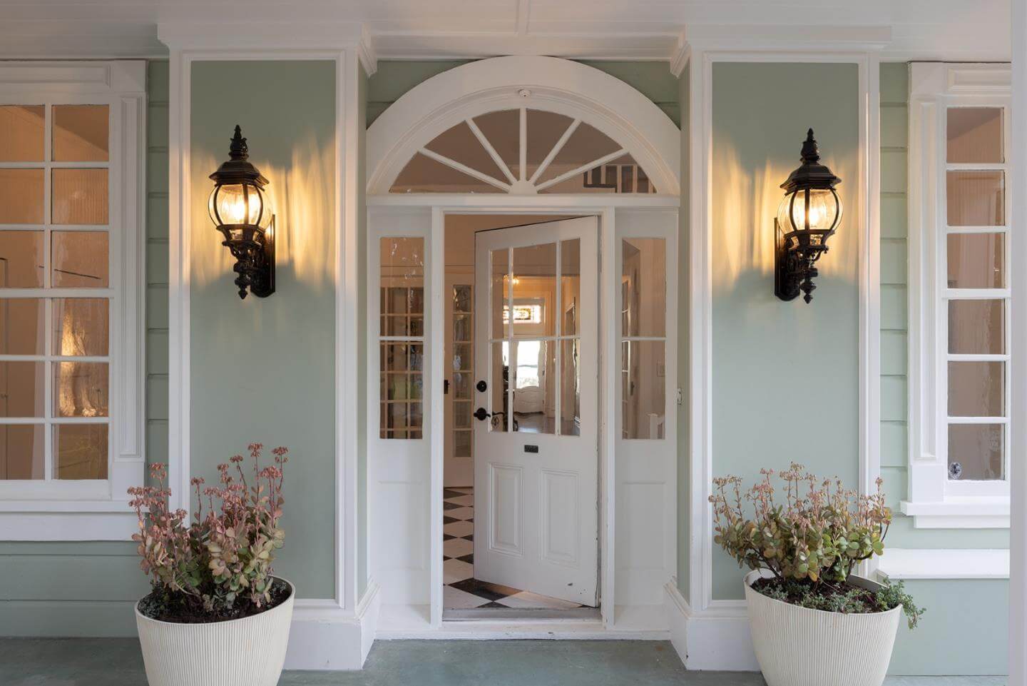 Santa Cruz Home Entryway With Classic Curb Appeal Close-up view of a Santa Cruz home entryway with a white front door, arched window detail, soft green exterior siding, decorative wall lanterns, and potted plants. This image highlights curb appeal, exterior presentation, and entryway features that can influence buyer perception and home value in Santa Cruz County.