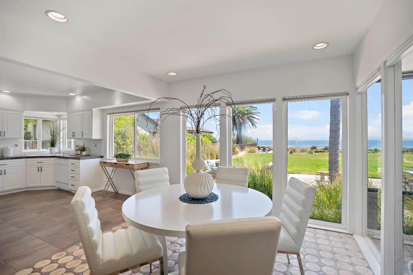 Santa Cruz Ocean View Dining Area With Bright Kitchen and Outdoor Views Bright Santa Cruz ocean view dining area with large windows, white kitchen cabinetry, natural light, tile flooring, and sliding glass doors overlooking a coastal yard and ocean view. This Santa Cruz home interior highlights the light, layout, kitchen connection, and outdoor space buyers often notice when comparing Santa Cruz properties.