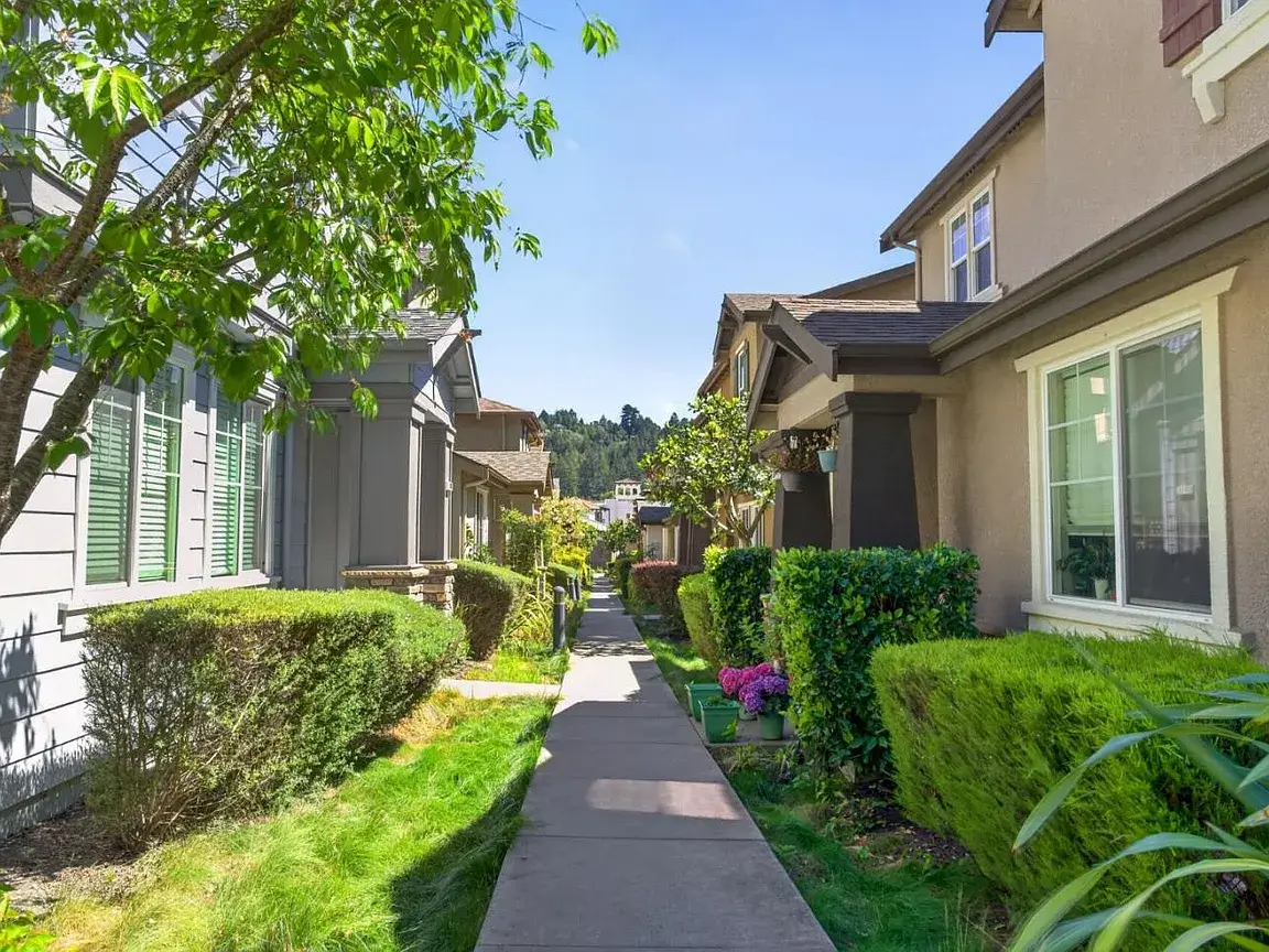 Santa Cruz Townhome Community With Landscaped Walkway Sunny exterior view of a Santa Cruz townhome community with a landscaped walkway, trimmed hedges, green lawns, mature trees, front windows, and well-maintained residential buildings. This image highlights curb appeal, neighborhood presentation, and property features that can matter during the Santa Cruz home selling timeline from listing preparation to buyer showings and closing.