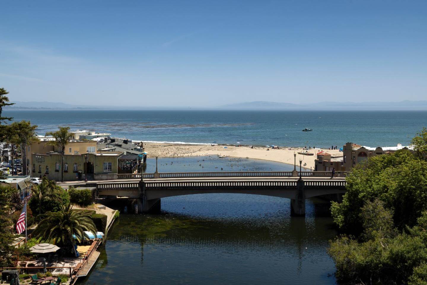 Scenic view of Capitola Beach in Santa Cruz County, with Soquel Creek flowing beneath a bridge toward the Monterey Bay, sandy shoreline, coastal buildings, and ocean views.