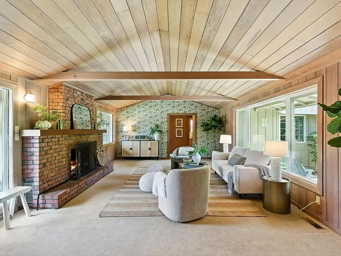 Warm living room in a Capitola-area home in Santa Cruz County, featuring a brick fireplace, exposed wood ceiling beams, large windows, wood-paneled walls, and a comfortable open seating area.