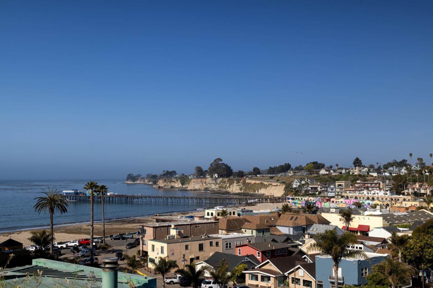 View of Capitola Village in Santa Cruz County, California, showing the Capitola Beach shoreline, historic pier, colorful beach cottages, palm trees, and coastal homes along the Monterey Bay waterfront.
